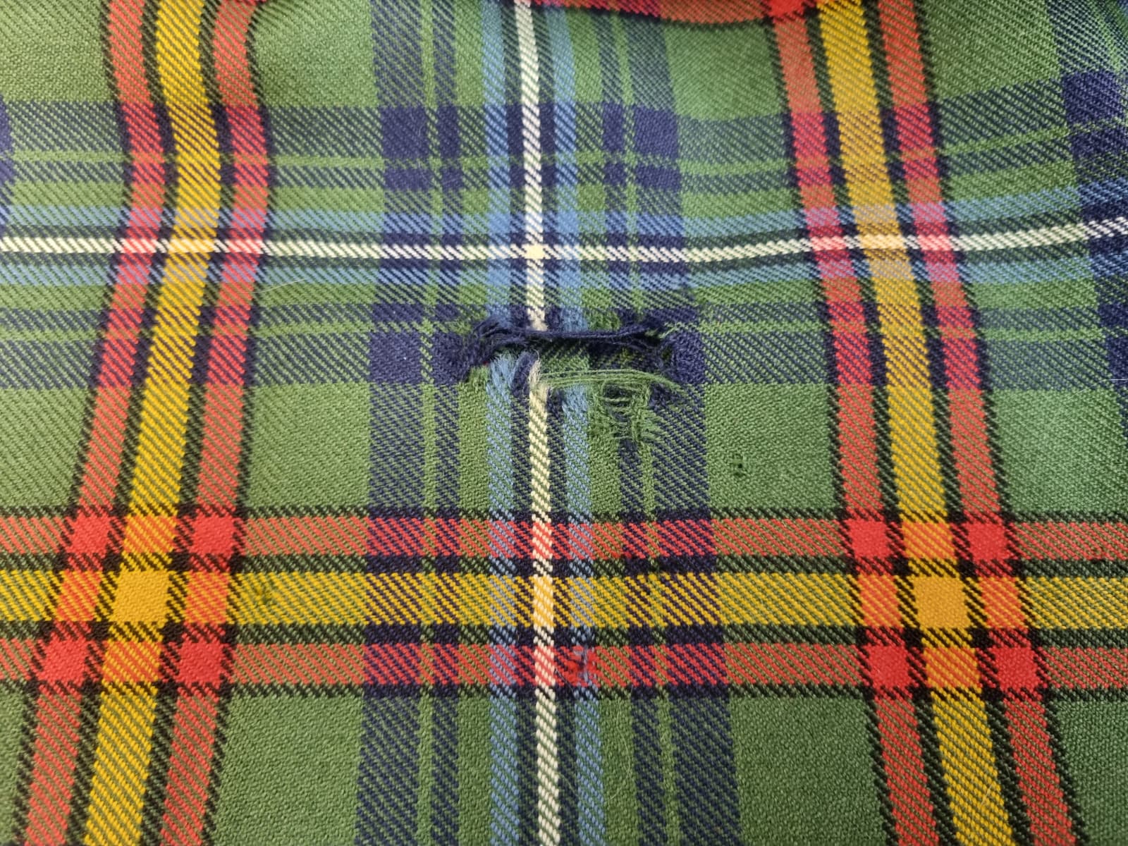 Green tartan kilt fabric showing a visible hole and broken fibres before repair.
